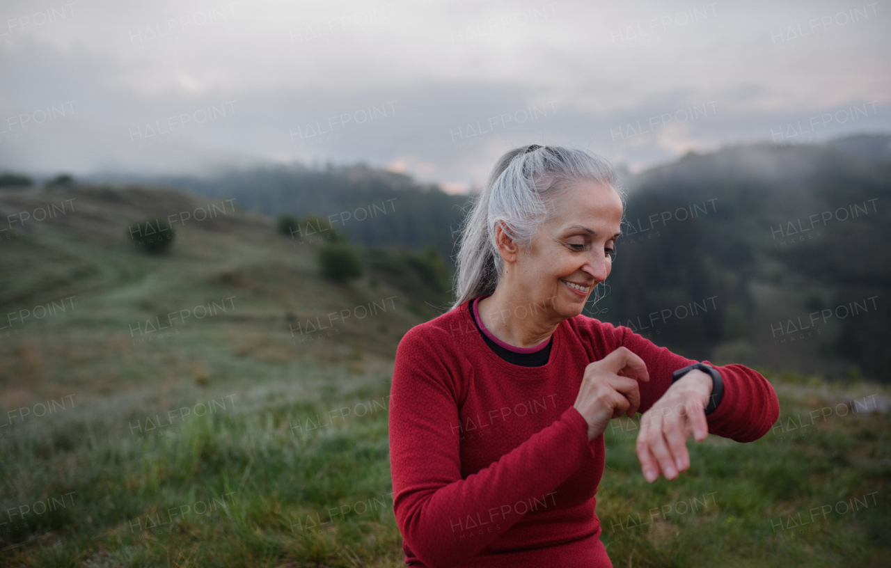 A senior woman jogger setting and looking at sports smartwatch, checking her performance in nature on early morning with fog and mountains in background.