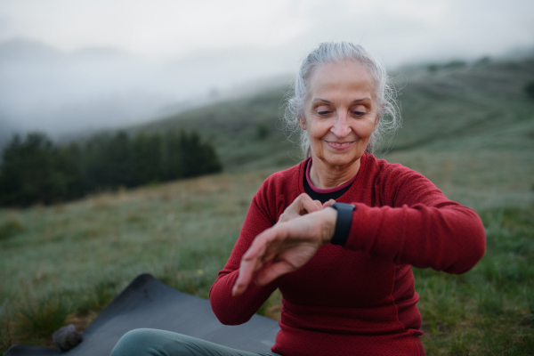 A senior woman jogger setting and looking at sports smartwatch, checking her performance in nature on early morning with fog and mountains in background.