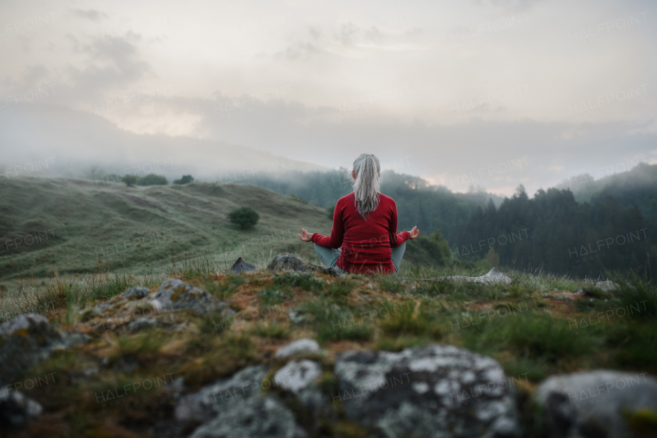 A rear view of senior woman doing breathing exercise in nature on early morning with fog and mountains in background.