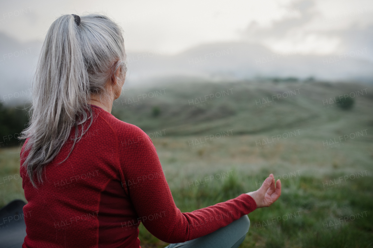 A rear view of senior woman doing breathing exercise in nature on early morning with fog and mountains in background.