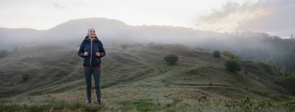 A senior woman jogging in nature on early morning with fog and mountains in background.