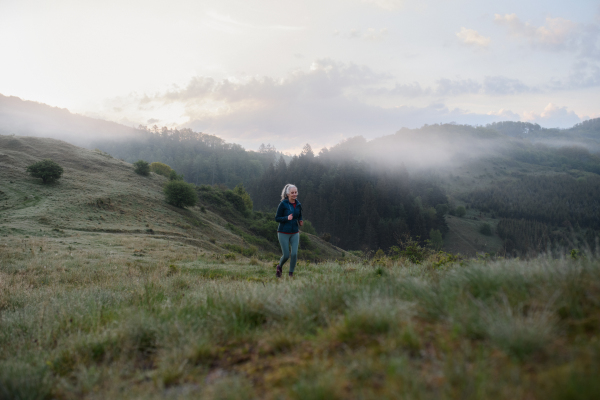 A senior woman jogging in nature on early morning with fog and mountains in background.