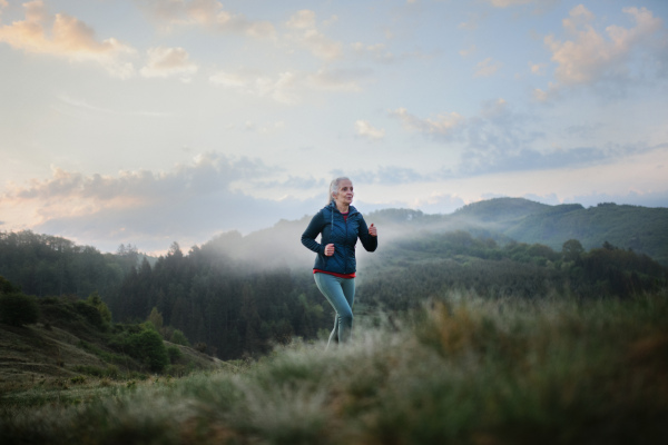 A senior woman jogging in nature on early morning with fog and mountains in background.