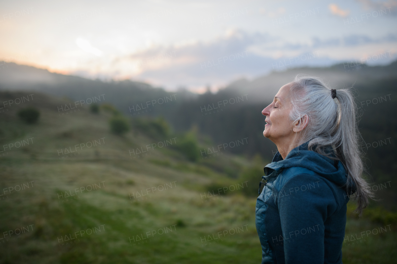 A senior woman doing breathing exercise in nature on early morning with fog and mountains in background.