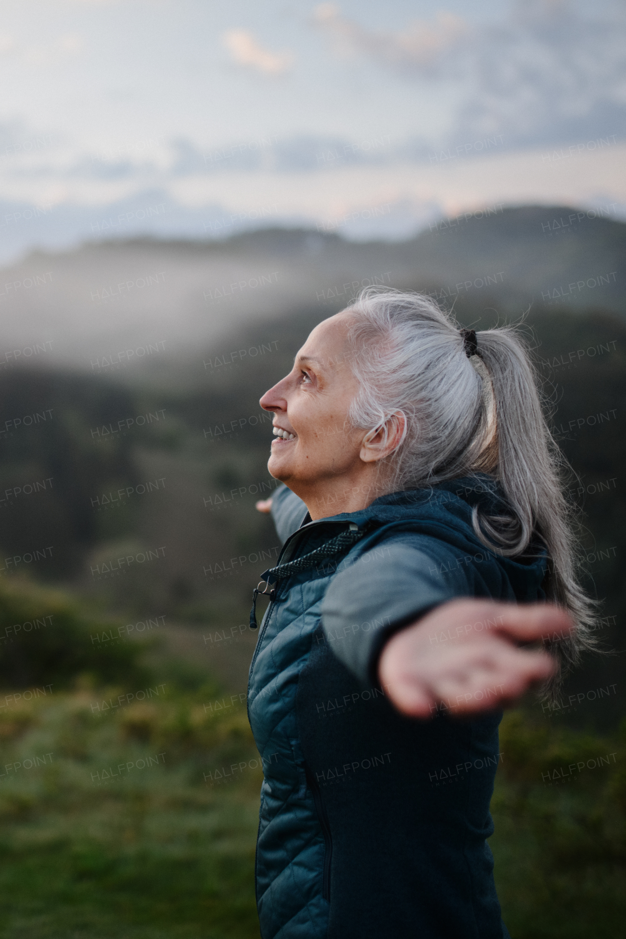 A senior woman doing breathing exercise in nature on early morning with fog and mountains in background.