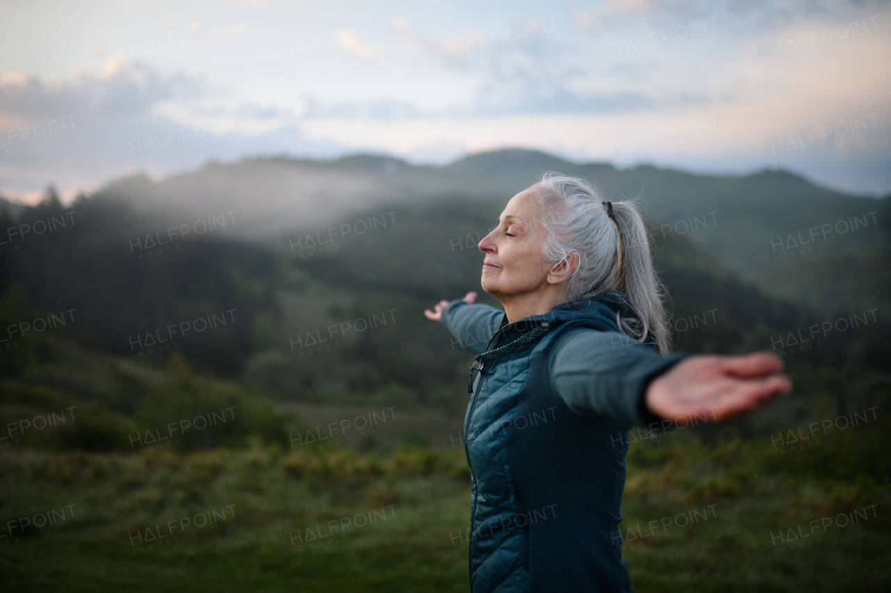 A senior woman doing breathing exercise in nature on early morning with fog and mountains in background.