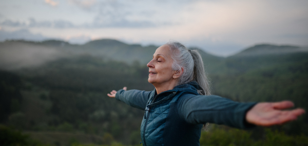 A senior woman doing breathing exercise in nature on early morning with fog and mountains in background.