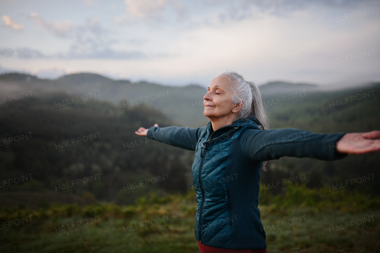 A senior woman doing breathing exercise in nature on early morning with fog and mountains in background.