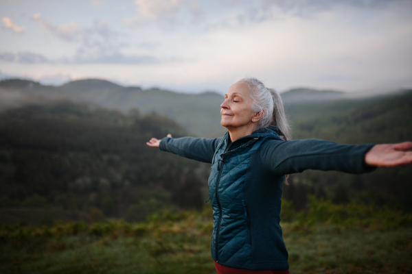 A senior woman doing breathing exercise in nature on early morning with fog and mountains in background.