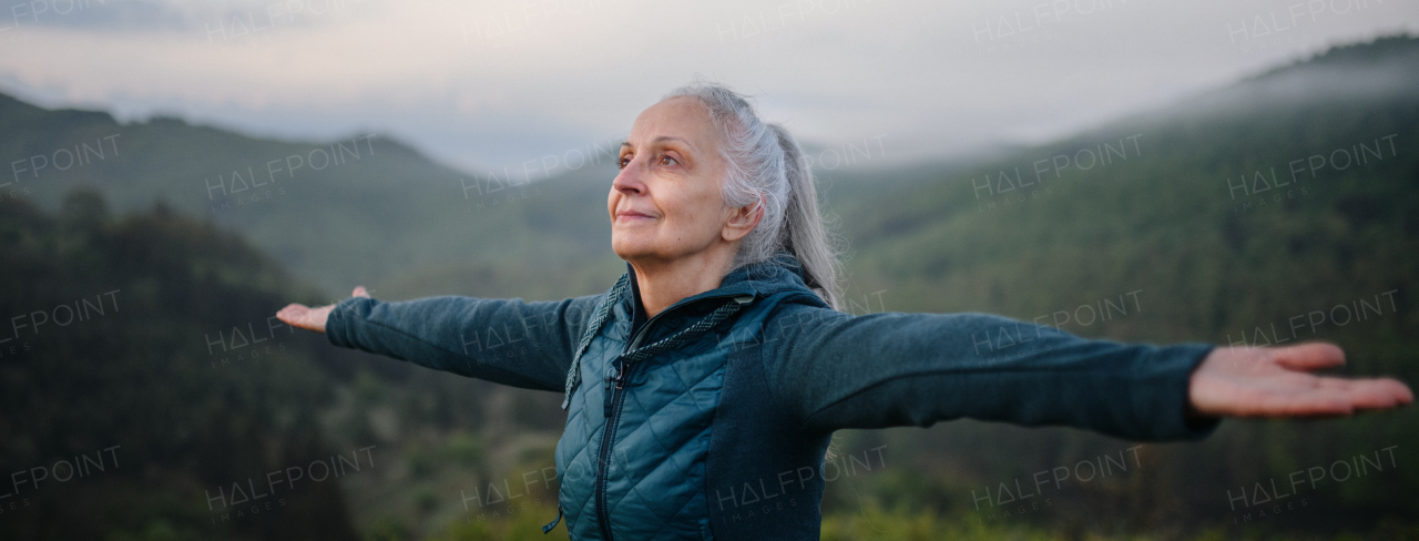 A senior woman doing breathing exercise in nature on early morning with fog and mountains in background.