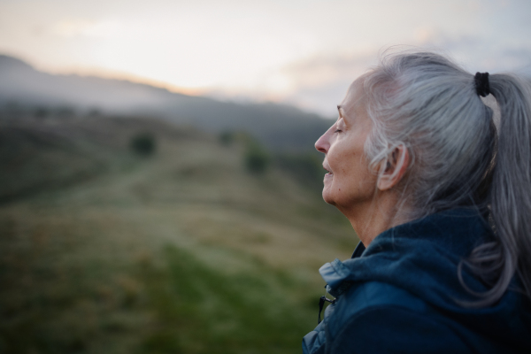 A senior woman doing breathing exercise in nature on early morning with fog and mountains in background.