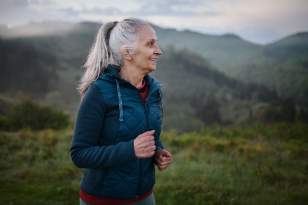 A senior woman jogging in nature on early morning with fog and mountains in background.