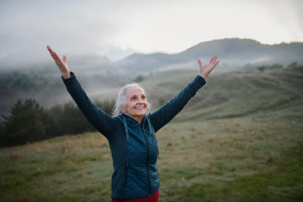 A senior woman doing breathing exercise in nature on early morning with fog and mountains in background.