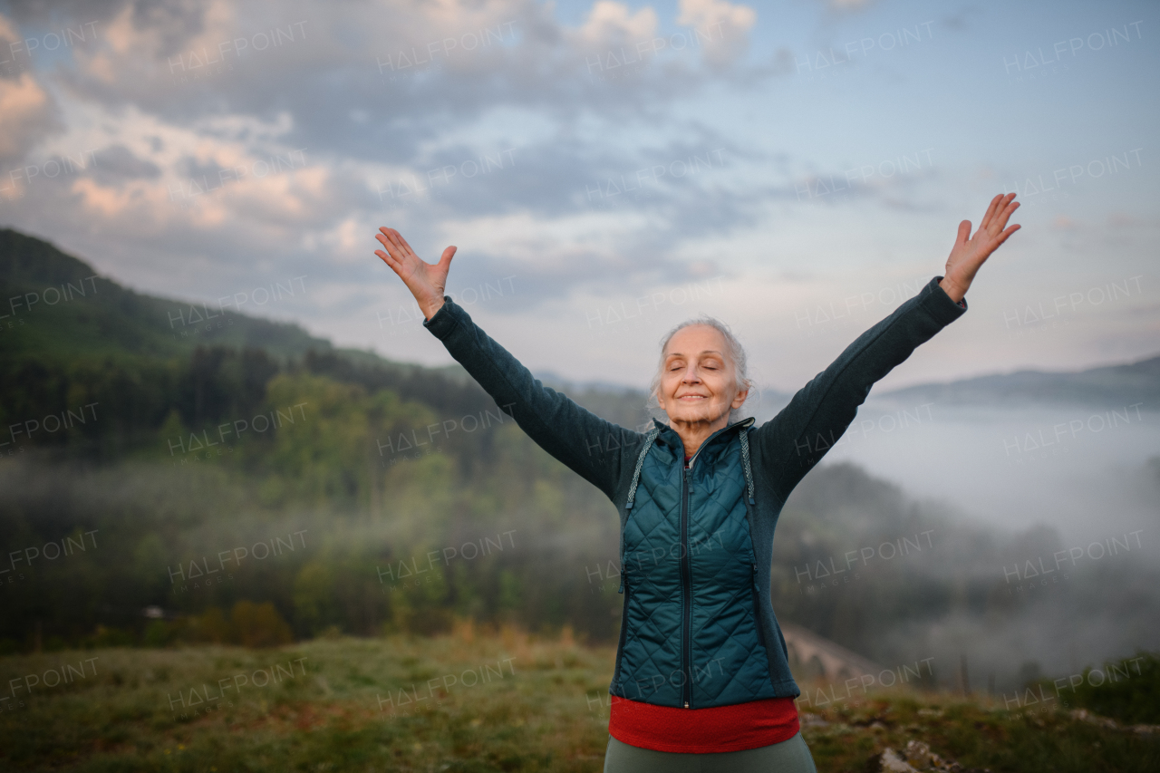 A senior woman doing breathing exercise in nature on early morning with fog and mountains in background.