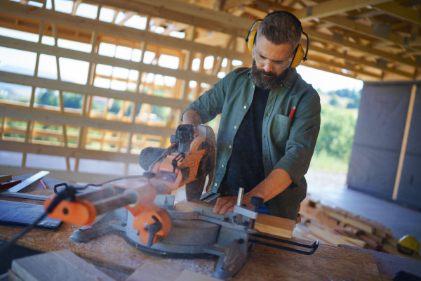 Construction worker working with the eletric saw inside wooden construction of house, diy eco-friendly homes concept.