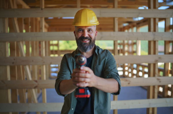 Construction worker posing with electric screwdriver in a wooden frame building, diy eco-friendly homes concept.