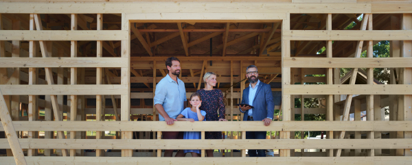 Sales agent showing unfinished ecologic wooden house to young family on construction site, and looking at view together.