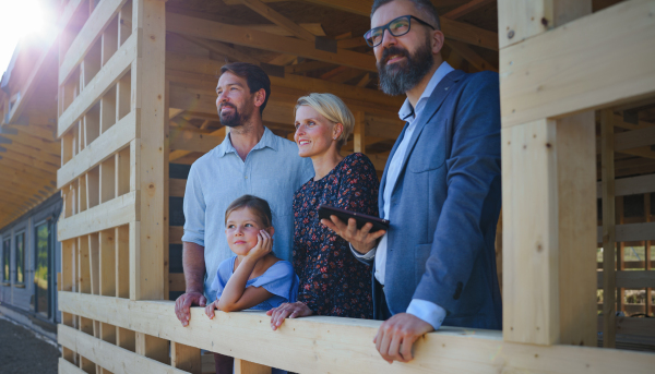 Sales agent showing the plans of new unfinished ecologic wooden house to young family on construction site, and looking at view together.