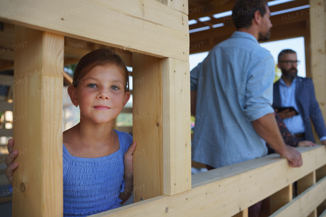 Little girl looking at camera, while her parents having meeting with house sales agent. Together in unfinished eco wooden house, concept of sustainiability and healthy living.