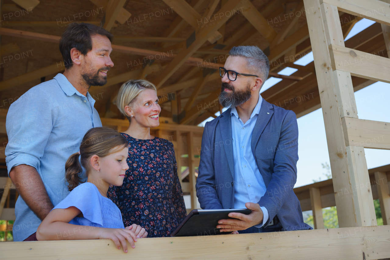 Sales agent showing the plans of new unfinished ecologic wooden house to young family on construction site, and looking at view together.