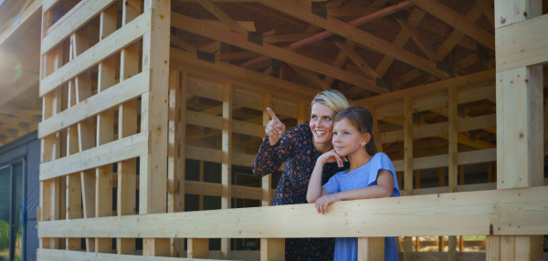 Mother and daughter on site inside of new ecology wooden home construction framing looking at view together.