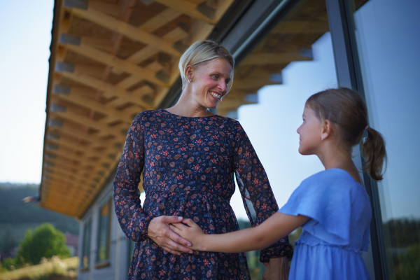 Little daughter stroking pregnant belly his mother, in front of their new modern wooden house.