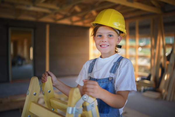 Happy little girl helping at their unfinished wooden house, standing at ladder with a helmet.