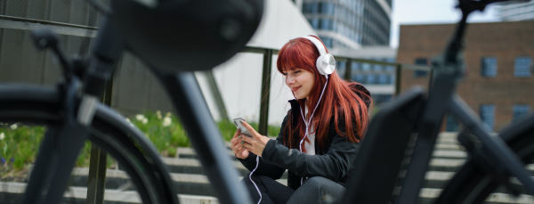 A portrait of businesswoman commuter on the way to work with bike, resting listening to music, sustainable lifestyle concept. Wide shot.