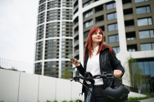 A portrait of businesswoman commuter on the way to work with bike, using smartphone, sustainable lifestyle concept.