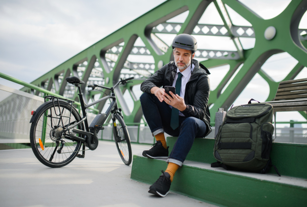 A businessman with bike sitting on bench, using smartphone. Commuting and alternative transport concept