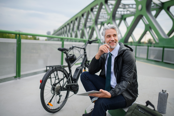 A businessman with bike sitting on bridge, using tablet and listening to music. Commuting and alternative transport concept