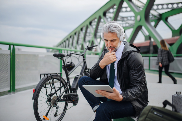 A businessman with bike sitting on bridge, using tablet and listening to music. Commuting and alternative transport concept