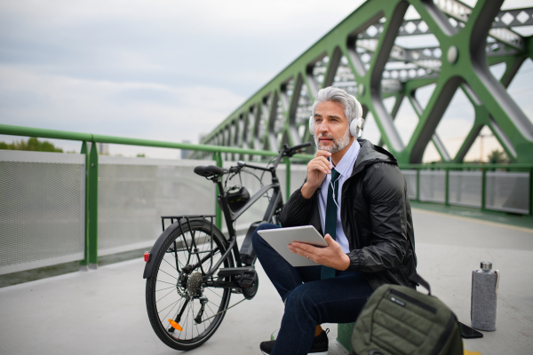 A businessman with bike sitting on bridge, using tablet and listening to music. Commuting and alternative transport concept