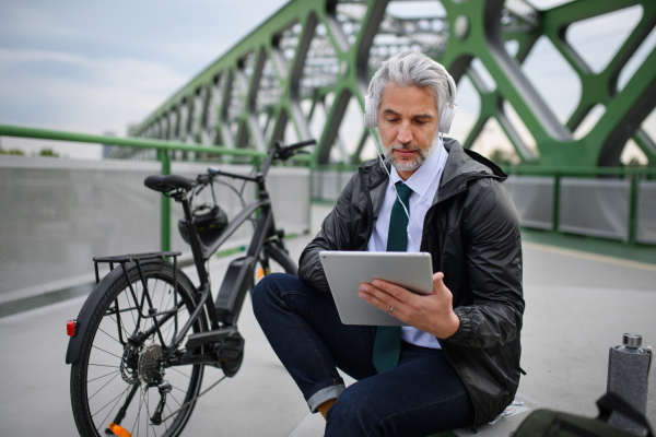 A businessman with bike sitting on bridge, using tablet and listening to music. Commuting and alternative transport concept