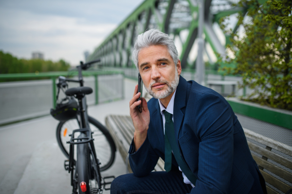 A businessman with bike sitting on bench, using smartphone. Commuting and alternative transport concept