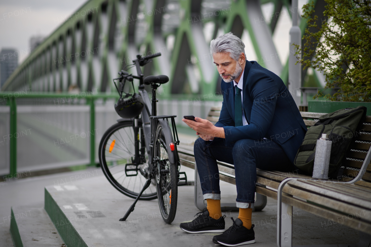 A businessman with bike sitting on bench, using smartphone. Commuting and alternative transport concept