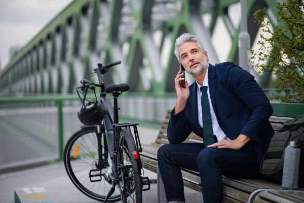 A businessman with bike sitting on bench, using smartphone. Commuting and alternative transport concept