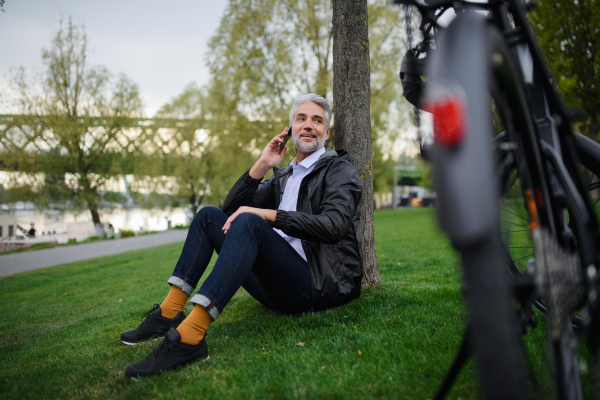 A businessman with bike sitting on grass in park, using smartphone. Commuting and alternative transport concept