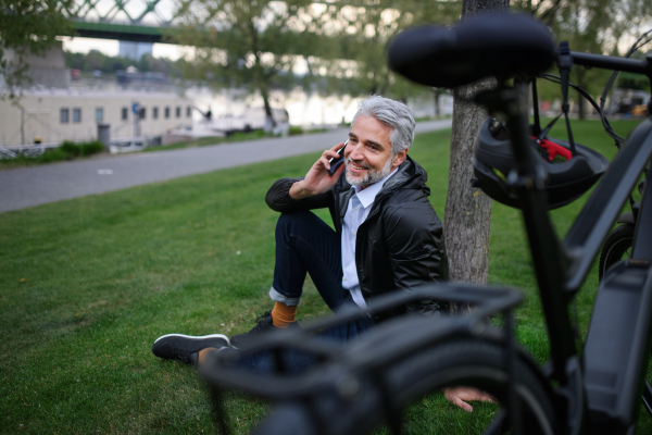 A businessman with bike sitting on grass in park, using smartphone. Commuting and alternative transport concept