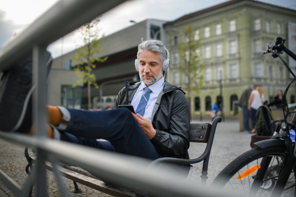 A businessman with bike sitting on bench, listening to music with feet up and resting. Commuting and alternative transport concept