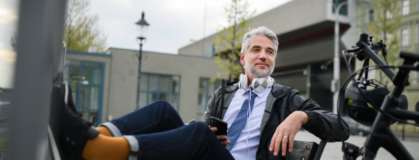 A businessman with bike sitting on bench, listening to music with feet up and resting. Commuting and alternative transport concept