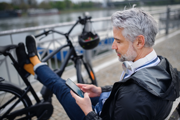 A businessman with bike sitting on bench, using smartphone. Commuting and alternative transport concept