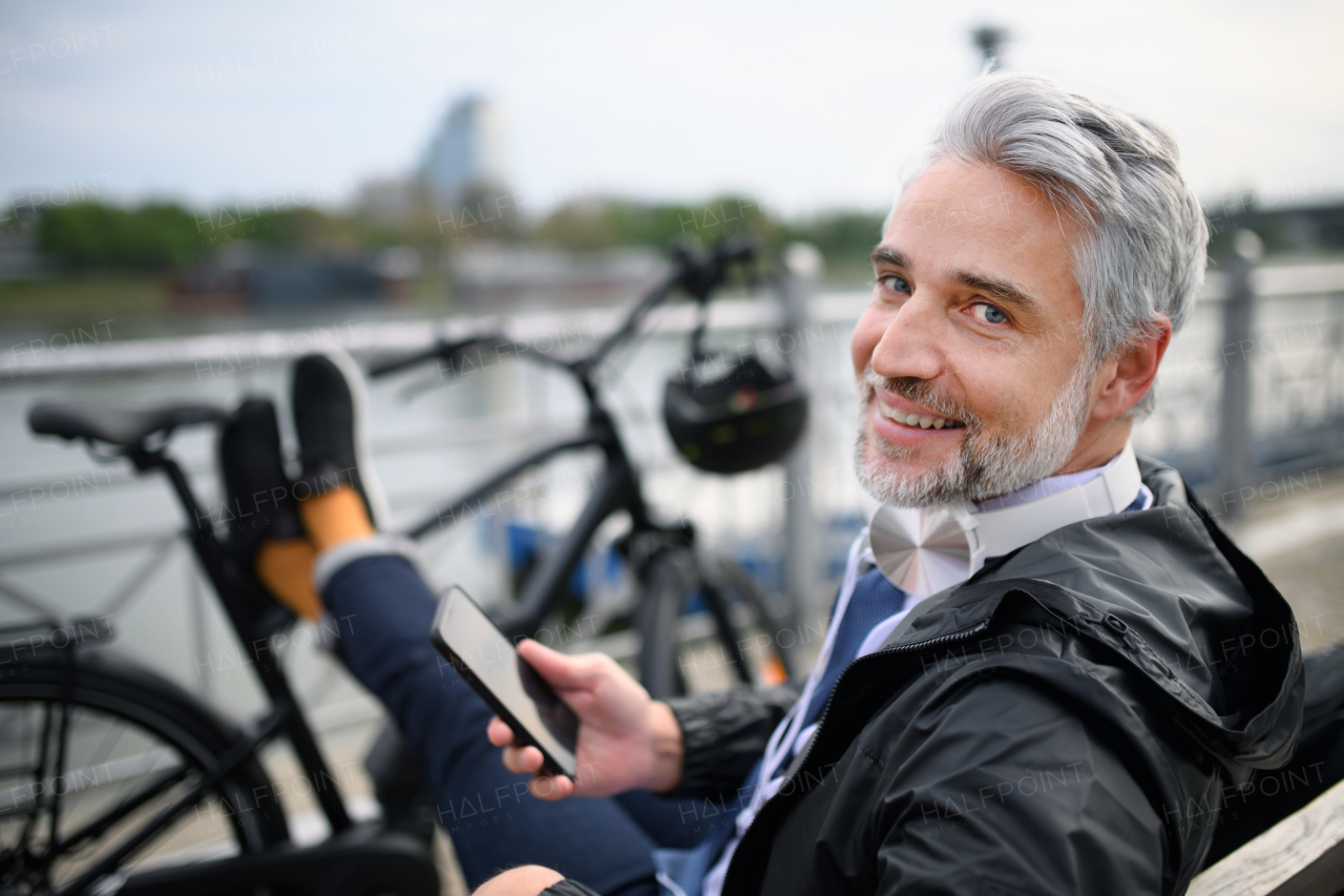 A businessman with bike sitting on bench, using smartphone. Commuting and alternative transport concept