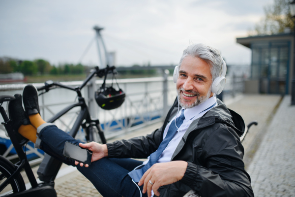 A businessman with bike sitting on bench, listening to music with feet up and resting. Commuting and alternative transport concept