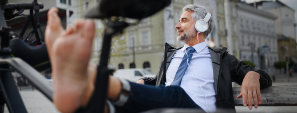 A businessman with bike sitting on bench, listening to music with feet up and resting. Commuting and alternative transport concept