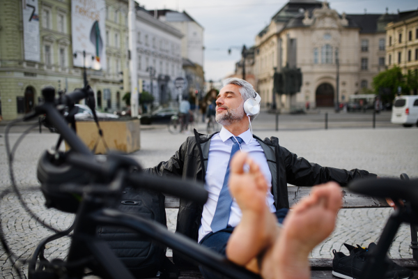 A businessman with bike sitting on bench, listening to music with feet up and resting. Commuting and alternative transport concept