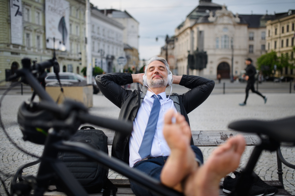 A businessman with bike sitting on bench, listening to music with feet up and resting. Commuting and alternative transport concept