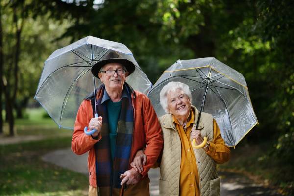 Happy senior couple walking with umbrellas in a park together.