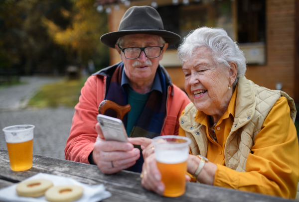 Happy senior couple in forest buffet resting after walk, having a beer and looking at smartphone.