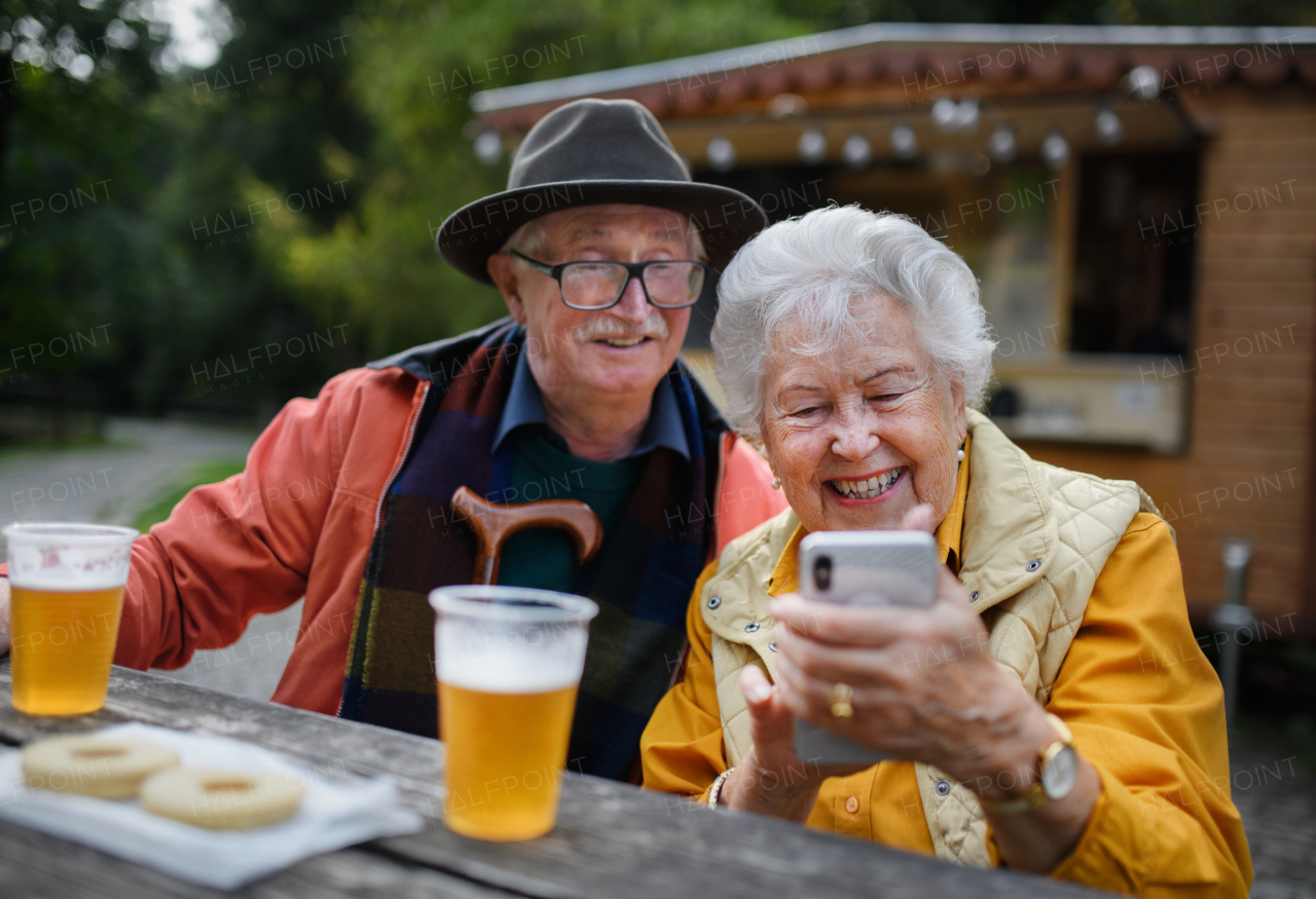 Happy senior couple in forest buffet resting after walk, having a beer and looking at smartphone.